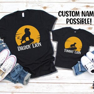 May include: Two black t-shirts with a yellow sun and a lion silhouette. The larger shirt says "Daddy Lion" and the smaller shirt says "Tommy Lion".  The shirts are laid out on a wood background with jeans and sneakers.
