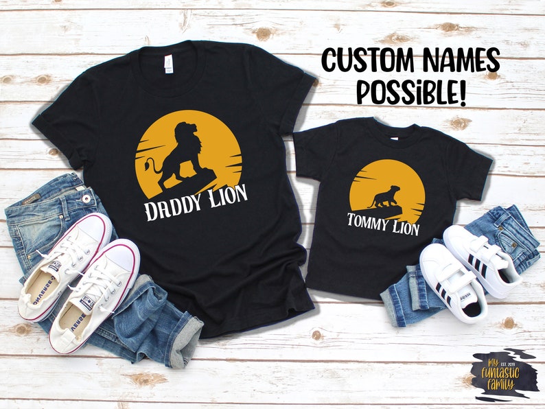 May include: Two black t-shirts with a yellow sun and a lion silhouette. The larger shirt says "Daddy Lion" and the smaller shirt says "Tommy Lion".  The shirts are laid out on a wood background with jeans and sneakers.