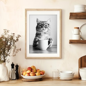 Op de afbeelding: Zwart-witfoto van een kat met een bril die een witte mok vasthoudt. De kat zit op een tafel en de foto is ingelijst in een zwart frame.