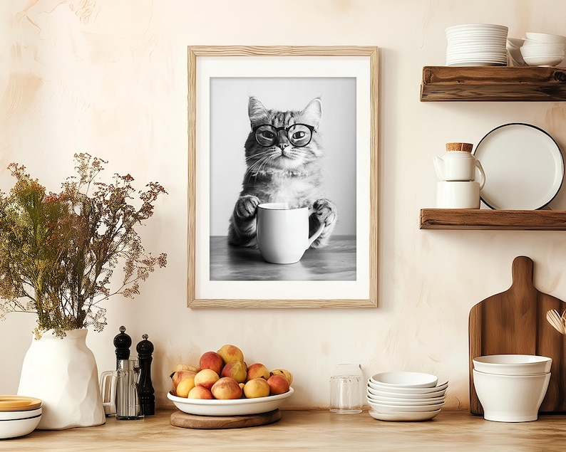 Op de afbeelding: Zwart-witfoto van een kat met een bril die een witte mok vasthoudt. De kat zit op een tafel en de foto is ingelijst in een zwart frame.