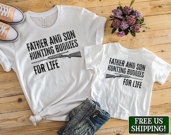 May include: Two white t-shirts with the text "Father and Son Hunting Buddies For Life" and a graphic of a rifle. The shirts are on a wooden surface.