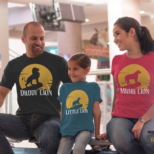 May include: A family of three wearing matching t-shirts with a lion silhouette and the words "Daddy Lion", "Mama Lion", and "Little Lion". The shirts are black, red, and teal. The family is standing in a shopping mall.