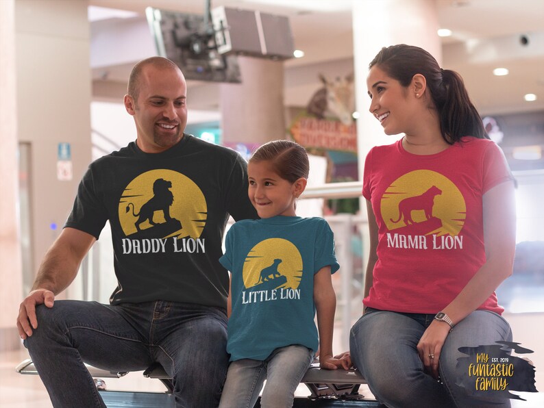 May include: A family of three wearing matching t-shirts with a lion silhouette and the words "Daddy Lion", "Mama Lion", and "Little Lion". The shirts are black, red, and teal. The family is standing in a shopping mall.