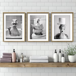 Op de afbeelding: Drie zwart-witte ingelijste prints van eenden in een badkamersetting. De eerste print toont een eend die zijn tanden poetst, de tweede toont een eend die een krant leest en de derde toont een eend die een toiletpapierhoed draagt.