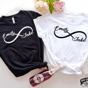 Op de afbeelding: Twee zwart-witte T-shirts met de namen "Emily" en "Jake" erop, verbonden door een oneindigheidssymbool. De shirts liggen op een wit oppervlak.