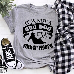 Peut inclure: Un t-shirt gris avec le texte "IT IS NOT A dad bod IT'S A father figure" imprimé dessus. Le texte est en noir et le t-shirt est posé à plat sur une surface blanche.