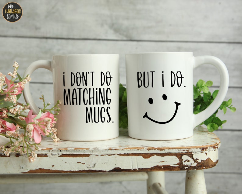 May include: Two white ceramic mugs with black text. One mug reads "I DON'T DO MATCHING MUGS." The other mug says "BUT I DO." with a smiling face. The mugs are on a distressed white wooden stool with pink flowers and greenery.