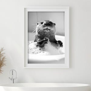 May include: A black and white photo of an otter in a bathtub full of bubbles. The otter is looking up at the camera with its paws on the edge of the tub.