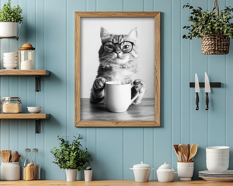 Op de afbeelding: Een zwart-witfoto van een kat met een bril die een witte mok vasthoudt. De kat zit op een houten tafel. De foto is ingelijst in een bruin houten lijst.