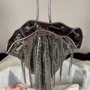 Könnte beinhalten: Eine silberne Mesh-Kordelzugtasche mit einem metallisch silbernen Griff. Die Tasche hat einen dunkel silbernen Rand und ist mit hängenden silbernen Fransen verziert. Die Tasche hängt vor einem weißen Hintergrund.