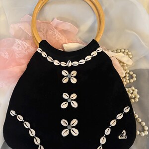 May include: Black handbag with a rounded shape and a light brown bamboo handle. The bag is decorated with white seashells and small, light blue accents. A small heart-shaped logo is visible on the lower right side.