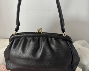 Puede incluir: Bolso de mano de cuero negro con diseño fruncido y cierre dorado. El bolso tiene una forma estructurada con un asa superior. El bolso está sostenido por una mano. El fondo incluye una tela rosa, perlas y guantes blancos.