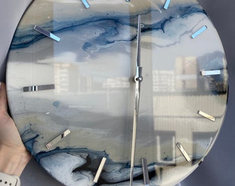 Puede incluir: Un reloj redondo blanco con vetas abstractas azules y grises. El reloj tiene manecillas plateadas y marcadores de hora plateados.