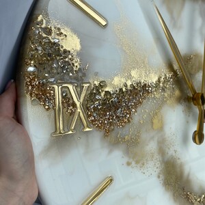 May include: A white and gold resin clock with Roman numeral markings and gold hands. The clock face has a marbled effect with gold and silver glitter embedded in the resin.