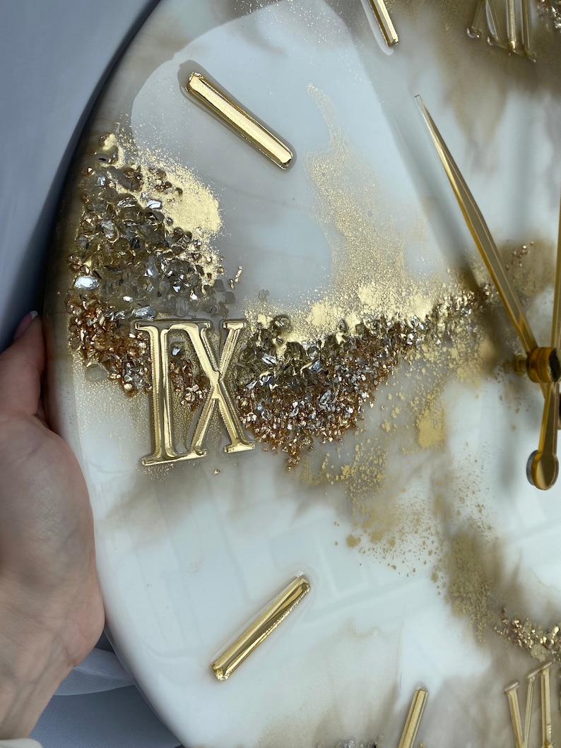 May include: A white and gold resin clock with Roman numeral markings and gold hands. The clock face has a marbled effect with gold and silver glitter embedded in the resin.
