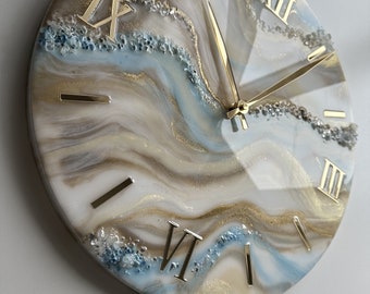May include: A round, white clock face with gold Roman numeral markings and gold hands. The clock face has a blue and brown abstract design with gold glitter accents.