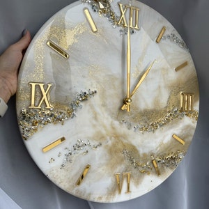 May include: A white and gold resin clock with Roman numeral markings and gold hands. The clock is 18 inches in diameter and has a glittery finish.