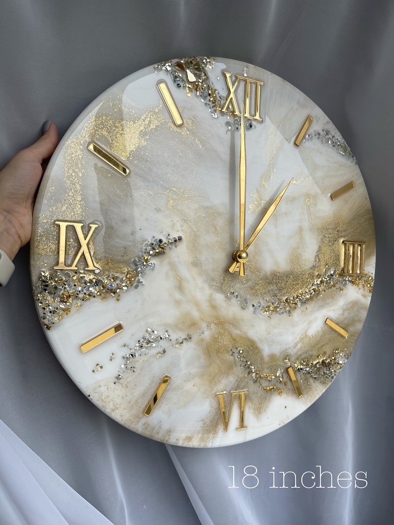 May include: A white and gold resin clock with Roman numeral markings and gold hands. The clock is 18 inches in diameter and has a glittery finish.