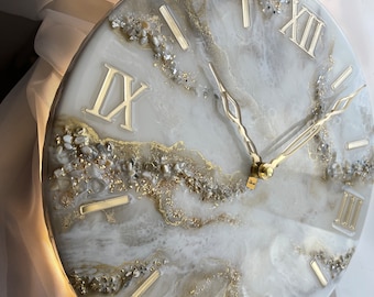 May include: White and gold resin clock with Roman numeral markings. The clock face has a marbled design with gold glitter and white and gold accents.