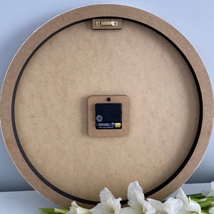 May include: The back of a round wooden clock with a black rim. The clock has a battery-operated quartz movement and a small white plastic hook for hanging.