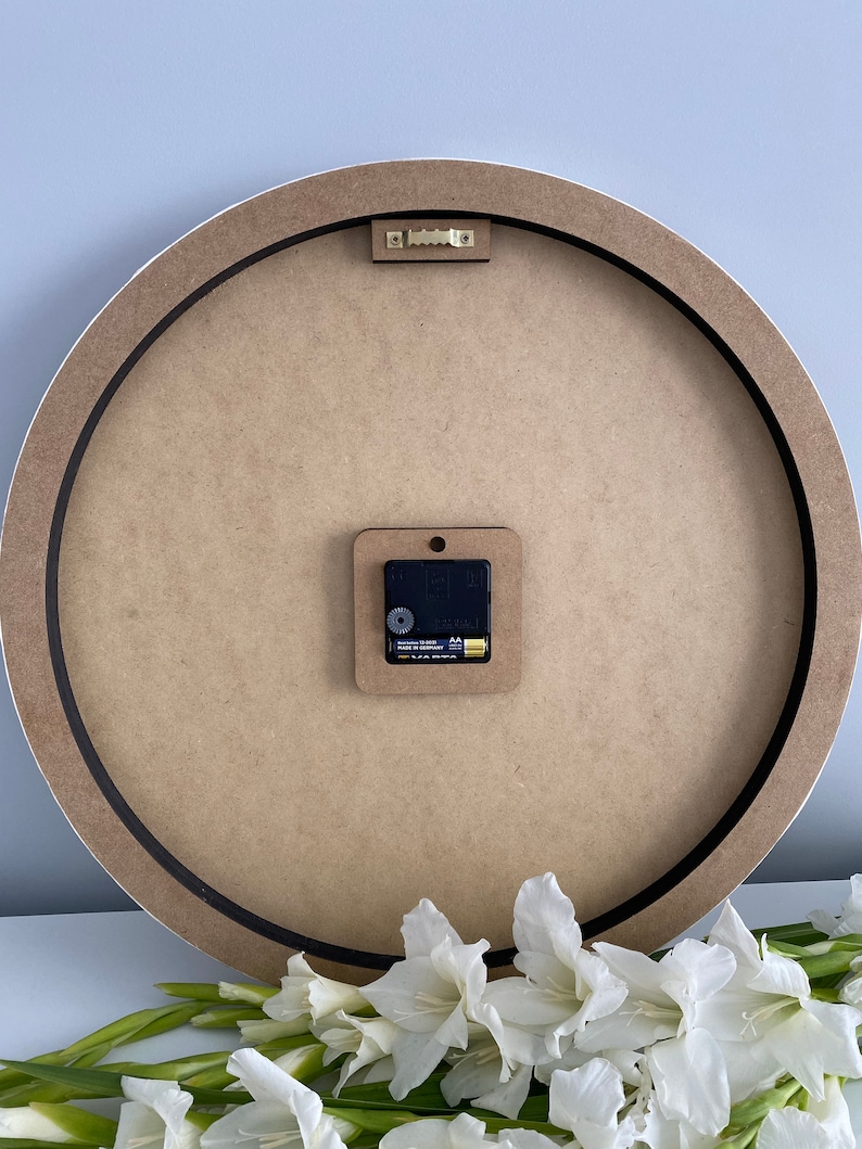 May include: The back of a round wooden clock with a black rim. The clock has a battery-operated quartz movement and a small white plastic hook for hanging.