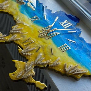 May include: A yellow and blue resin clock shaped like the outline of Ukraine with wheat stalks and Roman numerals. The clock face features a gold trident, the Ukrainian national symbol.