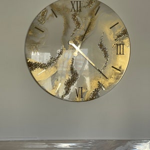 May include: A round, white and gold clock with Roman numeral markings and a glitter finish. The clock hands are black.
