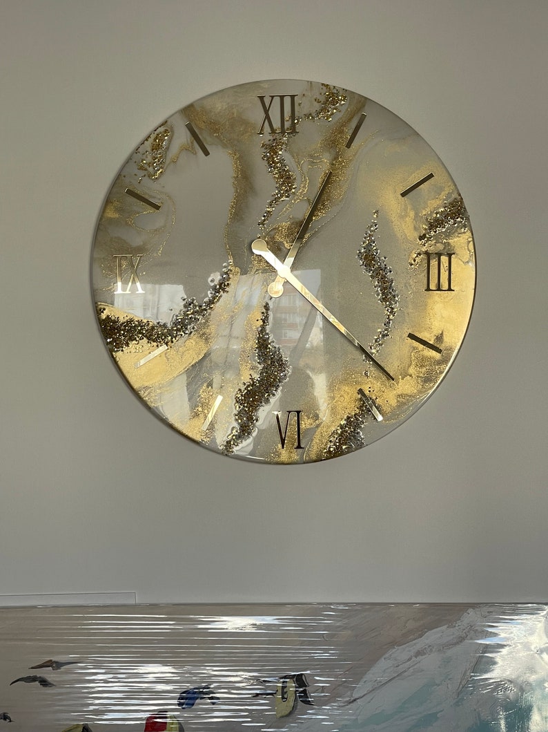 May include: A round, white and gold clock with Roman numeral markings and a glitter finish. The clock hands are black.