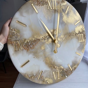 May include: White and gold resin clock with Roman numeral markings. The clock face has a marbled design with gold glitter accents.