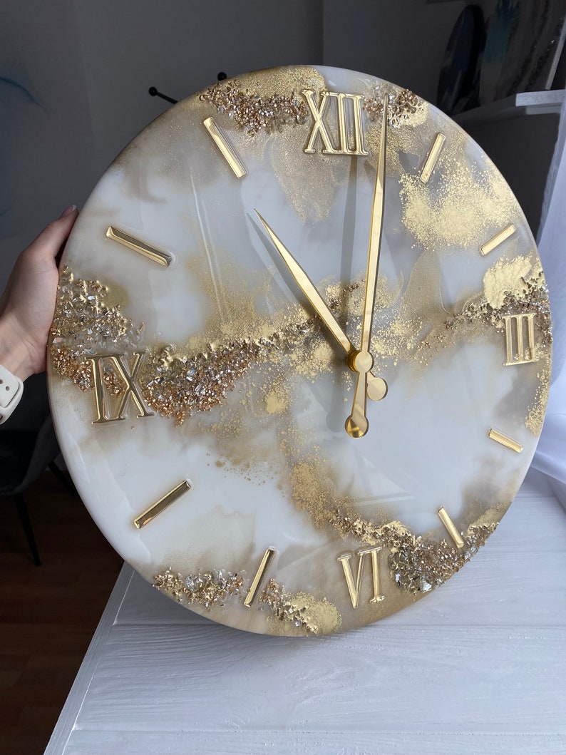 May include: White and gold resin clock with Roman numeral markings. The clock face has a marbled design with gold glitter accents.