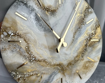 May include: Round wall clock with a white, gold, and gray marbled design. The clock face features gold-colored hands and hour markers. The design incorporates glitter and small stones for added texture and visual interest. The clock is a decorative home decor item.