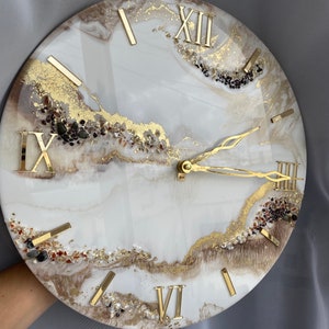 Puede incluir: Un reloj de resina blanco y marrón con números romanos dorados y manecillas doradas. La esfera del reloj tiene un diseño marmoleado con escamas doradas y pequeñas piedras blancas y marrones.
