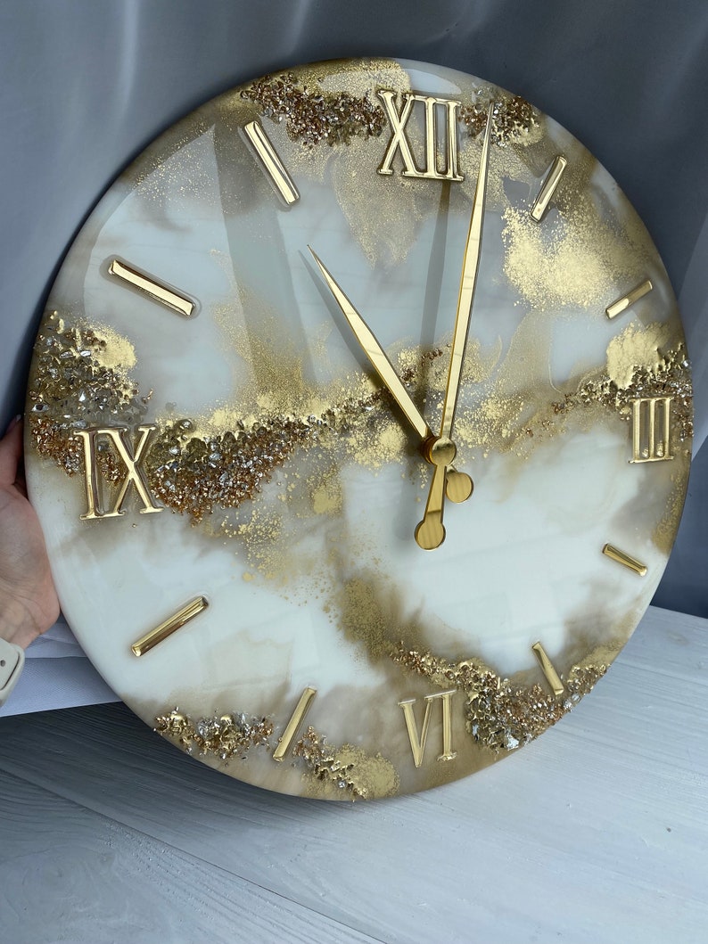 May include: A white and gold resin clock with Roman numeral markings. The clock face has a marbled effect with gold glitter embedded in the resin.