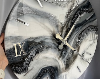 May include: A round wall clock with a marbled design in shades of white, gray, and black. The clock features silver Roman numerals and hands. Decorative crystals are embedded around the clock face, adding a touch of sparkle.