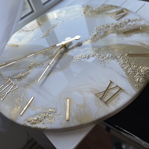 May include: A white and gold resin clock with Roman numeral markings. The clock face has a marbled design with gold glitter accents.
