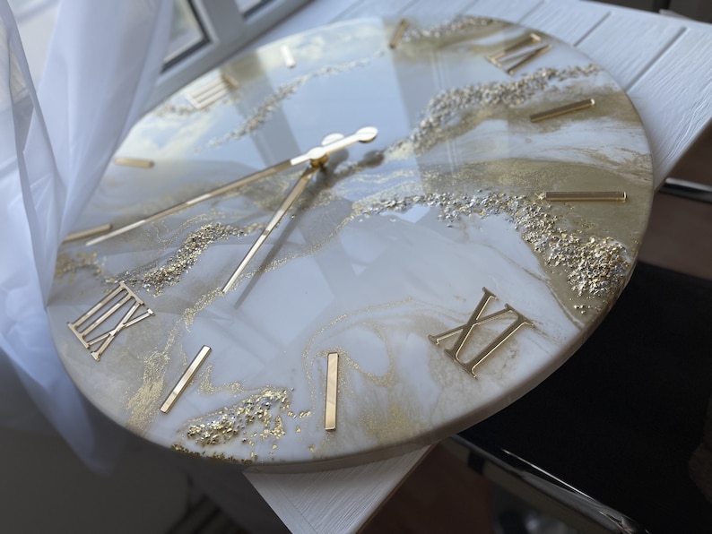 May include: A white and gold resin clock with Roman numeral markings. The clock face has a marbled design with gold glitter accents.
