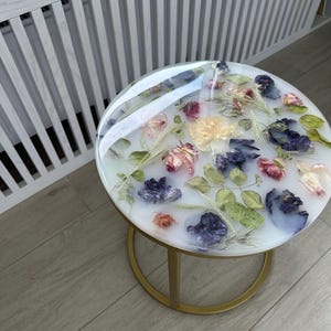 Op de afbeelding: Een ronde bijzettafel met een helder harsblad met daarin gedroogde bloemen in roze, paarse en gele tinten. De tafel heeft een goudkleurig metalen onderstel. De tafel heeft een diameter van ongeveer 46 cm.