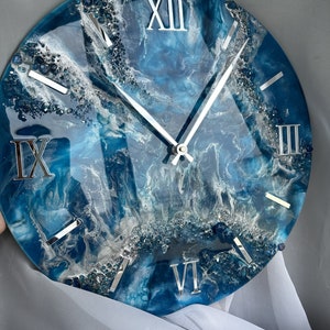 Può includere: Un orologio in resina blu e bianca con numeri romani. Il quadrante dell'orologio ha un design astratto a spirale con glitter argentato incorporato nella resina.