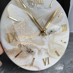 Op de afbeelding: Witte en gouden harsklok met Romeinse cijfers. De wijzerplaat heeft een marmerachtig ontwerp met gouden accenten.