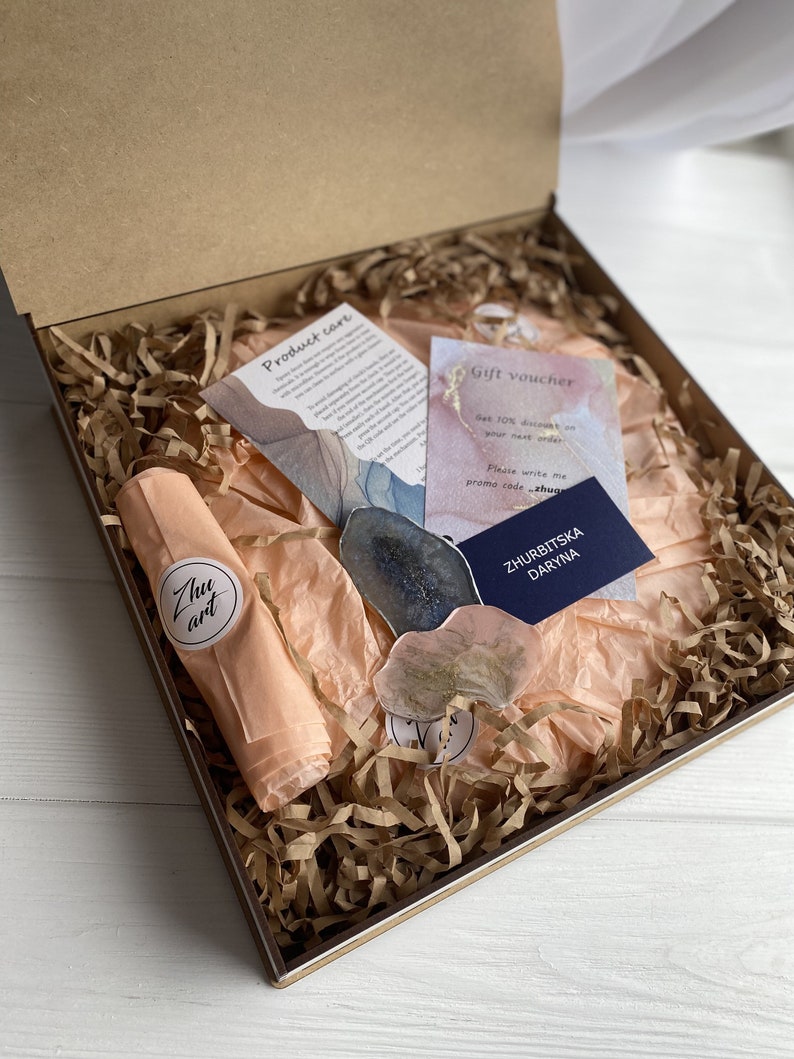 May include: A gift box filled with brown packing paper and a peach-colored tissue paper. The box contains two resin art pieces, a gift voucher with the text "Get 10% discount on your next order. Please write me promo code: zhug", and a business card with the text "ZHURBITSKA DARYNA".