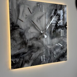 May include: Square wall clock with a marbled black and white design. The clock face features silver hands and Roman numerals. The clock has a glowing, warm-toned border, adding a modern touch to the decor.