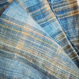 Op de afbeelding: Close-up van een geweven stof in blauw en geel met een textuurpatroon. De stof is gevouwen en toont de ingewikkelde details van de weefstructuur.