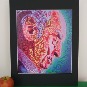 Op de afbeelding: Een kleurrijke portretprint van een profiel van een persoon, met een levendige mix van rode, roze, gele en blauwe tinten. Het kunstwerk is omlijst met een zwart passe-partout en een witte rand. Een appel rust op het groene oppervlak eronder.