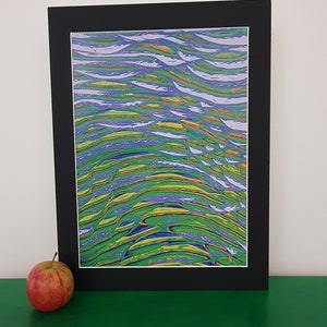 Op de afbeelding: Abstracte kunstafdruk met een zwarte passe-partout. Het kunstwerk toont vloeiende lijnen in tinten groen, blauw, geel en oranje, waardoor een golvend patroon ontstaat. Een rood-gele appel ligt links van het ingelijste kunstwerk op een groen oppervlak.