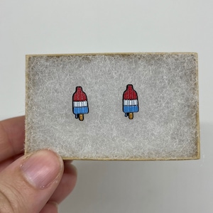 Pode incluir: Um par de brincos de botão de picolé vermelho, branco e azul em uma pequena caixa branca e marrom.