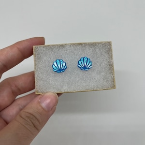 May include: A pair of blue and white seashell stud earrings in a small white box. The seashells have a glittery finish.