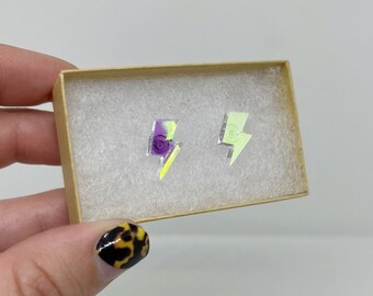 May include: A pair of lightning bolt shaped earrings in a small cardboard box. One earring is yellow and green, the other is purple and green.