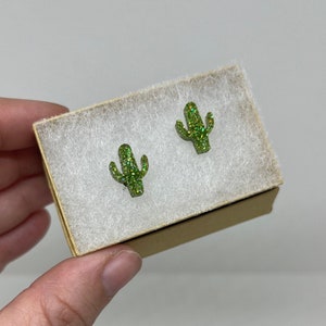 May include: A pair of green glitter cactus earrings in a small white box with a brown lid.
