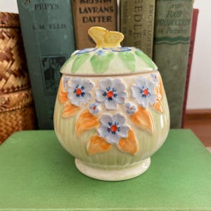 May include: A ceramic jar with a lid, decorated with blue floral designs and orange leaves on a light green base. The lid has green leaves and a yellow handle. The jar is placed in front of a stack of vintage books.