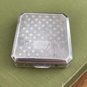 May include: A vintage silver Stratton compact with a geometric pattern of dots and stars. The compact has a rectangular shape with rounded corners and a small clasp. The text "Stratton Made in England" is visible on the top of the compact.